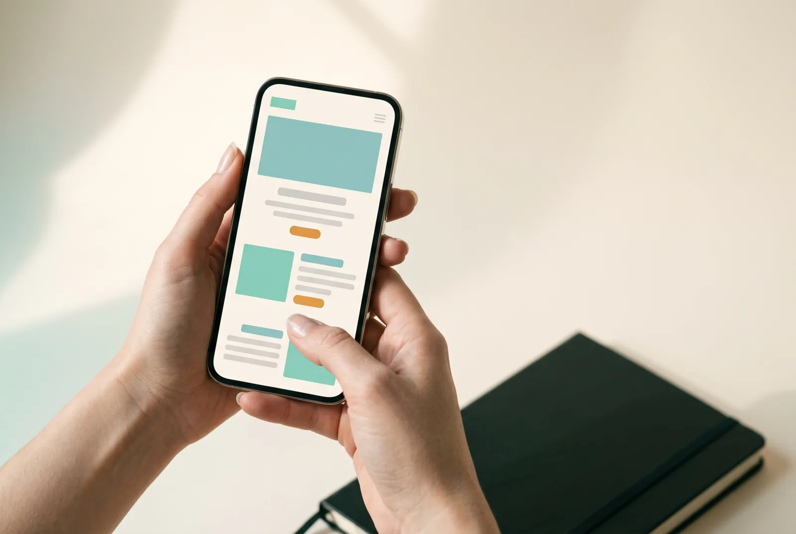 Hands holding a phone showing a simple mobile website layout in teal and amber, with a dark notebook on a cream desk behind