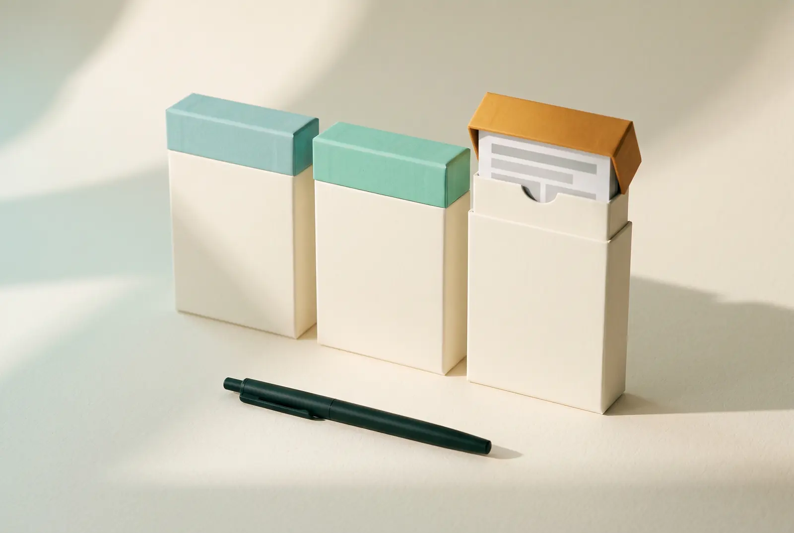 Three identical cream boxes standing in a row with lids in pale teal, mint and amber, the amber one open just enough to show a layout card inside, beside a dark pen